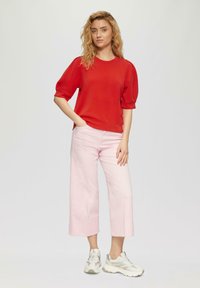 Top rosso a maniche corte con scollatura tonda, abbinato a jeans a gamba larga rosa chiaro e sneakers bianche, su uno sfondo neutro.