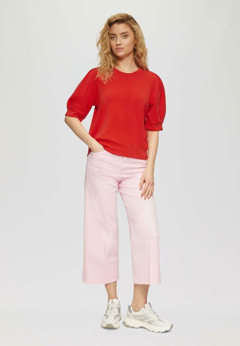 Top rosso a maniche corte con scollatura tonda, abbinato a jeans a gamba larga rosa chiaro e sneakers bianche, su uno sfondo neutro.