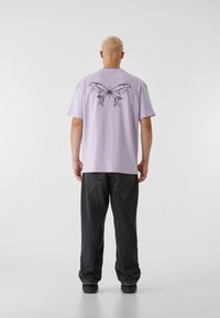 Lavendel kortermet t-skjorte med et svart sommerfugl-design på baksiden. Sammen med løstsittende svarte jeans og mørke sko.