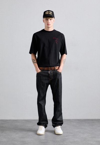 Jeune homme portant une casquette noire, un t-shirt noir avec du texte rouge, un jean sombre avec une ceinture marron, des baskets blanches, se tenant devant un fond gris clair uniformément.