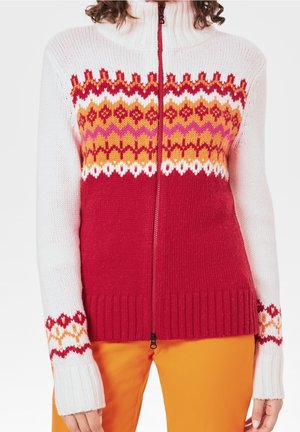 Kvinde iført en hvid og rød strikket lynlås-sweater med orange og pink mønstret bærestykke og orange bukser, vist fra midt på kroppen og ned.