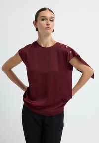 Blusa bordeaux con maniche corte, tessuto liscio, scollatura rotonda, dettagli con bottoni sulle spalle, abbinata a pantaloni neri.