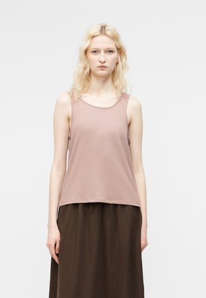 Mladá žena s blond vlasmi, oblečená v svetlom fialovom topom bez rukávov a tmavohnedej sukni, stojaca pred jednoduchým bielym pozadím.