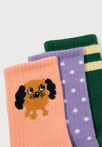 Três meias coloridas: uma pêssego com um cão de desenho animado, uma roxa com bolinhas brancas e uma verde com listras amarelas, empilhadas sobre um fundo branco.
