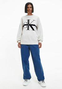 Sudadera de Calvin Klein Jeans, color crema, corte oversize, con estampado de logo en negro. Combinada con jeans de mezclilla azul de pierna ancha y zapatillas blancas.