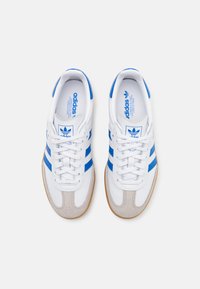Hvite sneakers med blå striper og subtil grå semsket skinn. Har snøring og gummisåle for godt grep.