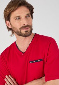 Rode T-shirt met korte mouwen en V-hals van lichtgewicht materiaal, met een marineblauwe zak waarin accenten van ankerpatroon op de zak en hals zijn verwerkt.