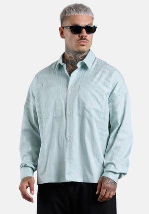 Mann mit kurzen grauen Haaren und Hals-Tattoos, der schwarze Sonnenbrillen und ein hellblaues gestreiftes Hemd mit Knöpfen trägt.