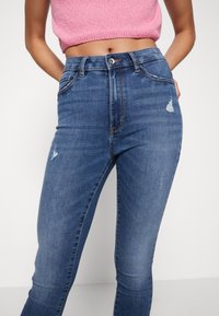 Mujer vestida con jeans skinny azules de tiro alto con ligeras desgastaduras y un top de punto rosa corto, con las manos en las caderas.