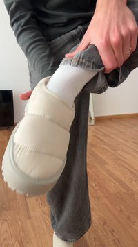 Zapato sin cordones acolchado color crema, usado con calcetín blanco. Costura en la parte superior y punta redondeada. Persona ajustando unos pantalones de mezclilla mientras está sentado en el suelo de madera.