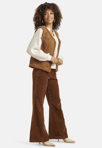 Brown faux shearling vest over a white blouse with embroidered sleeves, paired with high-waisted brown corduroy pants and beige heels.