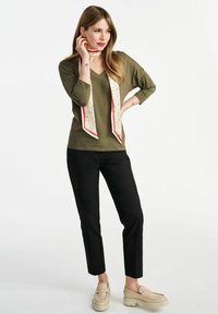 Zielono-oliwkowa bluzka z dekoltem w serek, czarne zwężane spodnie, beżowe platformowe loafersy oraz wzorzysta kremowa chusta z czerwoną lamówką na szyi.