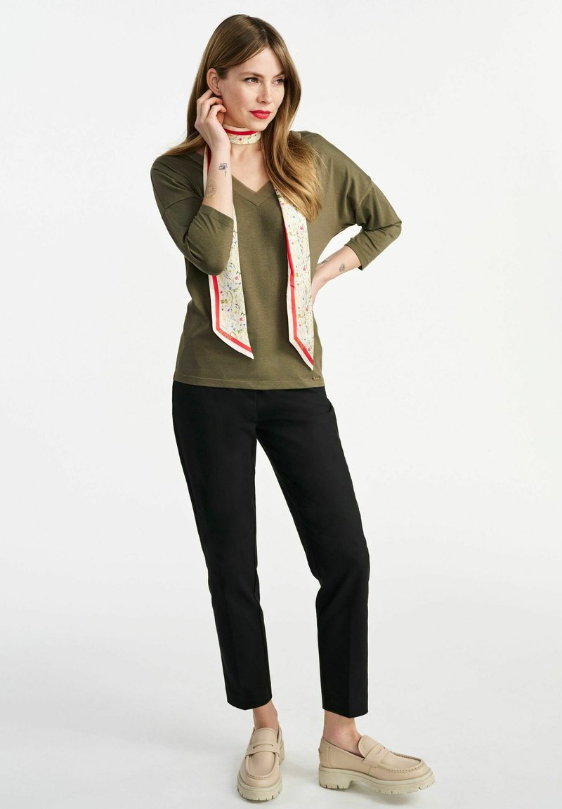 Olijfgroen V-hals top, zwarte tapered broek, beige platform loafers en een gemusterde crèmekleurige sjaal met rode rand om de nek.