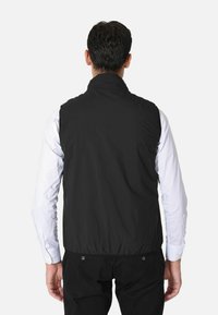 Gilet nero senza maniche realizzato in tessuto liscio e leggero con colletto alto e orlo elastico. Indossato sopra una camicia bianca a maniche lunghe.