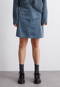 Falda en línea A de corduroy en azul suave, con un cierre frontal de botones, bolsillos laterales y un dobladillo que cae por encima de la rodilla. Combinada con zapatos negros.