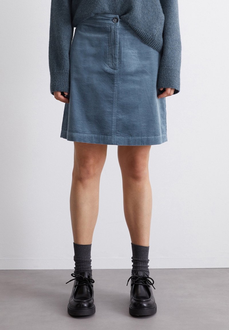 Falda en línea A de corduroy en azul suave, con un cierre frontal de botones, bolsillos laterales y un dobladillo que cae por encima de la rodilla. Combinada con zapatos negros.