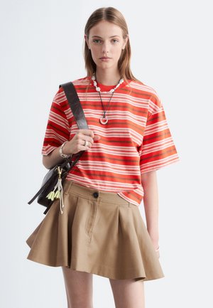 Jeune femme portant un t-shirt rayé rouge et blanc, une jupe plissée beige, un collier, un bracelet et un sac à bandoulière noir avec des pompons.