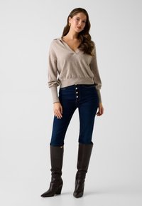 Beżowy sweter z dzianiny z dekoltem w serek i długimi rękawami, w połączeniu z ciemnoniebieskimi jeansami z wysokim stanem, zapinanymi na duże złote guziki, oraz brązowymi, sięgającymi kolan kozakami.