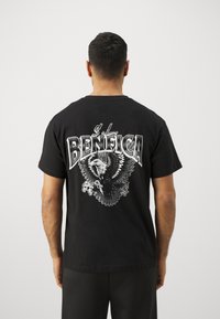 Melnas kokvilnas T-krekls ar lielu baltu un pelēku ērgļa grafiku un tekstu "S.L. BENFICA" aizmugurē. Īsās piedurknes un apaļais izgriezums.