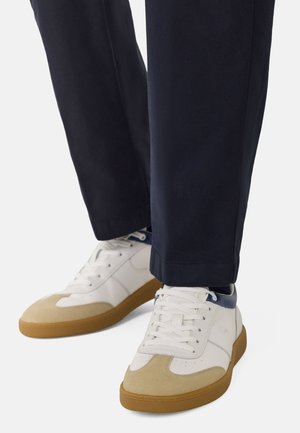 Witte sneakers met beige suède neuzen, blauwe accenten en rubberen gumzolen, gecombineerd met navy broek voor een casual uitstraling.