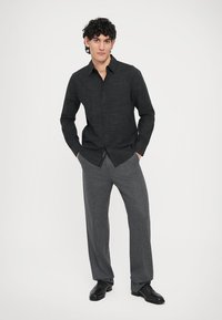 Sort skjorte med knapper og et diskret ternet mønster, kombineret med grå dressbukser. Modellen står med hænderne i lommerne og har sorte sko på.