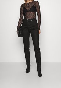 Czarny koronkowy body z długim rękawem nałożone na czarny biustonosz, w połączeniu z czarnymi spodniami z wysokim stanem i czarnymi botkami na szpilce.