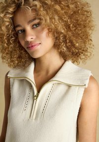 Strick-Ärmelloses Top in Creme mit einem Reißverschlusskragen, ausgestattet mit gerippten Kanten, zarten Musterdetails und einer glatten Textur.