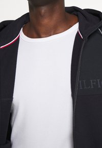 Sudadera negra con cremallera y ribetes rojos y blancos en la capucha; camiseta gris debajo; tela suave y texturizada; marca sutil en el lateral.