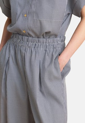 Personne portant une chemise à rayures verticales bleu et blanc et un pantalon assorti avec une ceinture élastique, main dans la poche droite, boutons beige sur la chemise.