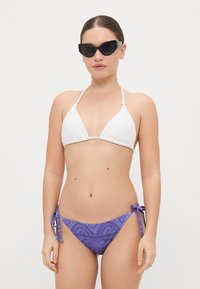 Biely textúrovaný trojuholníkový bikiny top s nastaviteľnými ramienkami v kombinácii s fialovými vzorovanými spodnými bikiny s viazaním na bokoch. Čierne slnečné okuliare v tvare mačacích očí.