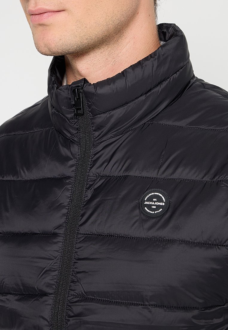 Svart vadderad jacka med hög krage, dragkedja och strukturerad yta. Har en cirkulär logotyp på vänster bröstområde.