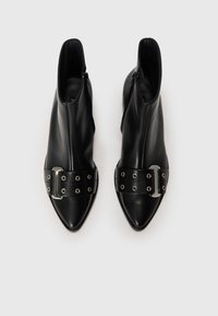 Botas de tornozelo em couro preto com bicos pontudos, apresentando uma larga faixa preta com olhais prateados e um detalhe de fivela metálica. Fecho com zíper.