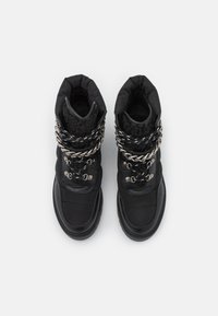 Par de botas de invierno negras con cuellos acolchados, cordones en blanco y negro, detalle de cadena metálica y tela texturizada sobre un fondo blanco.