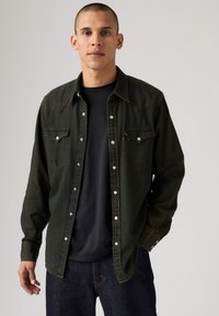 Camicetta in denim verde scuro con bottoni a pressione, due tasche sul petto e colletto classico. Presenta cuciture a contrasto e una vestibilità comoda.