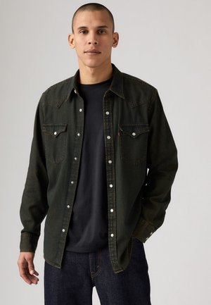 Camisa de mezclilla verde oscuro con botones a presión, dos bolsillos en el pecho y un cuello clásico. Presenta costuras en contraste y un ajuste relajado.