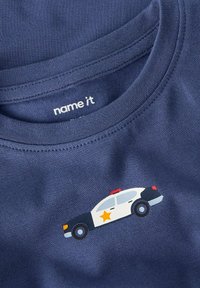 Primo piano del colletto di una camicia blu con una piccola grafica di una macchina della polizia che presenta una stella dorata e una luce rossa nella zona del petto.