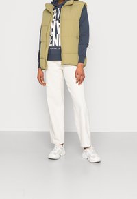 Olivgrön pufferväst över en marinblå hoodie, i kombination med vita vida byxor och ljusgrå sneakers. Slät textur, dragkedjestängning.