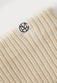 Cream-colored textile with a ribbed texture, featuring a black logo tag. Material is soft with a structured design.