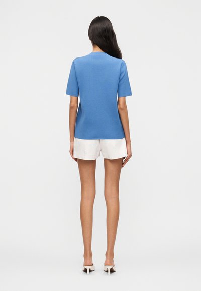 Γυναίκα με μακριά σκούρα μαλλιά στέκεται με την πλάτη προς τα εμπρός φορώντας μπλε μπλούζα με κοντά μανίκια, λευκά σορτς και λευκά πέδιλα με τακούνι σε λευκό φόντο.