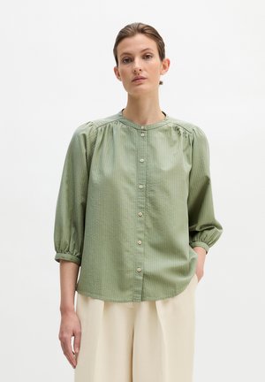 Femme portant un chemisier vert clair boutonné avec des manches trois-quarts bouffantes et un pantalon large crème, debout devant un fond blanc uni.