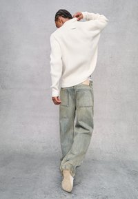 Pullover crema a coste con colletto alto, abbinato a jeans larghi blu chiaro con due tasche posteriori e leggere sfumature. Sneakers bianche visibili.