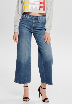 Nainen, joka käyttää vyötäröltä korkeavyötäröisiä, leveälahkeisia sinisiä farkkuja, lyhytvartista harmaata collegepaitaa ja mustia nilkkaremmiä sisältäviä korkokenkiä.