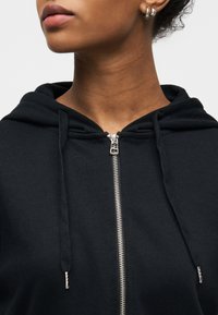 Gros plan sur une personne portant un sweat à capuche noir avec une fermeture éclair argentée et des cordons, montrant le bas du visage et le cou.