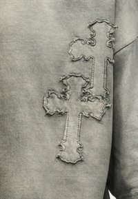 Γκρίζο ύφασμα με δύο ραμμένες μπαλώματα σε σχήμα σταυρού με φθαρμένες άκρες στην περιοχή του στήθους ενός ενδύματος.