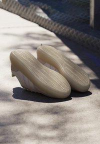 Beige träningsskor med texturerade gummisålar, släta läderöverdelar och tre vertikala blå ränder på sidorna.