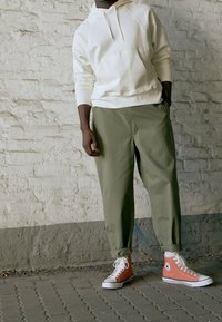 Bílá mikina s kapucí a přední kapsou, spojena s olivově zelenými zúženými kalhotami a oranžovými vysokými teniskami s bílou podrážkou.