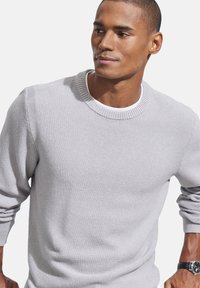 Hellgrauer Strickpullover mit rundem Halsausschnitt, gerippten Bündchen und strukturiertem Finish. Über einem weißen T-Shirt getragen.