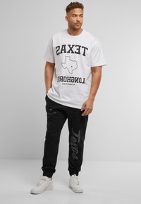 Hvid T-shirt med sort "TEXAS LONGHORNS" grafik og Texas kontur; parret med sorte joggers med subtil grå "Texas" tekst og hvide sneakers.