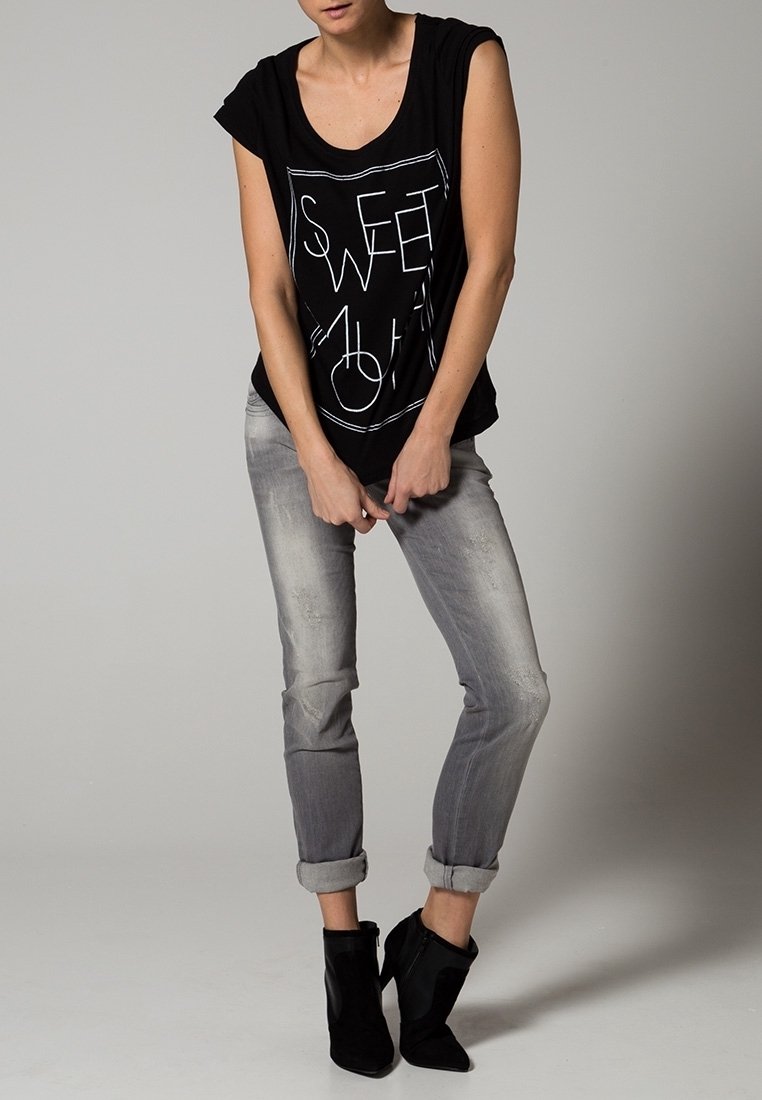 Černé bezrukávové tričko s bílým geometrickým potiskem se slovy „SWEET.“ V kombinaci se světle šedými džíny s ohrnutými nohavicemi a černými kotníkovými botami.