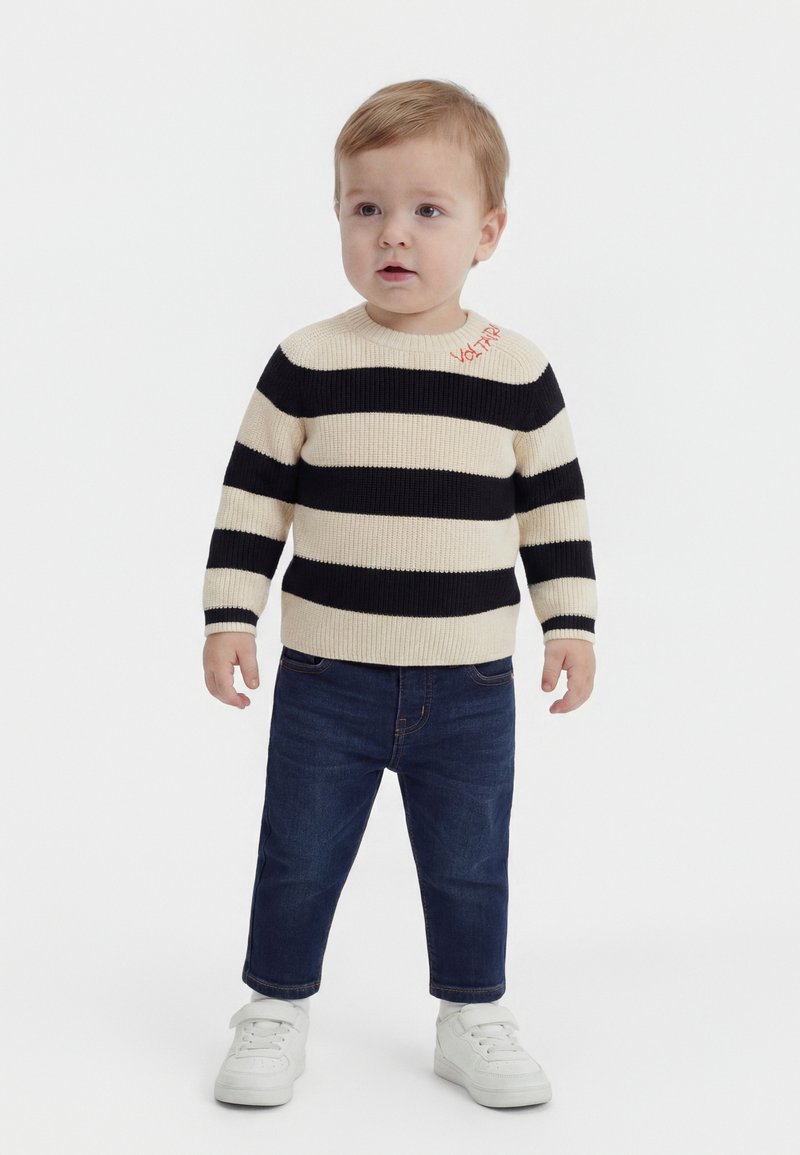 Νήπιο με ανοιχτό καστανά μαλλιά που φοράει μαύρο-άσπρο ριγέ πουλόβερ, σκούρα μπλε τζιν και λευκά αθλητικά παπούτσια, στέκεται σε λευκό φόντο.